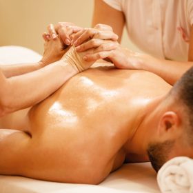 Female therapists massaging man's back while holding hands during spa treatment at wellness center.